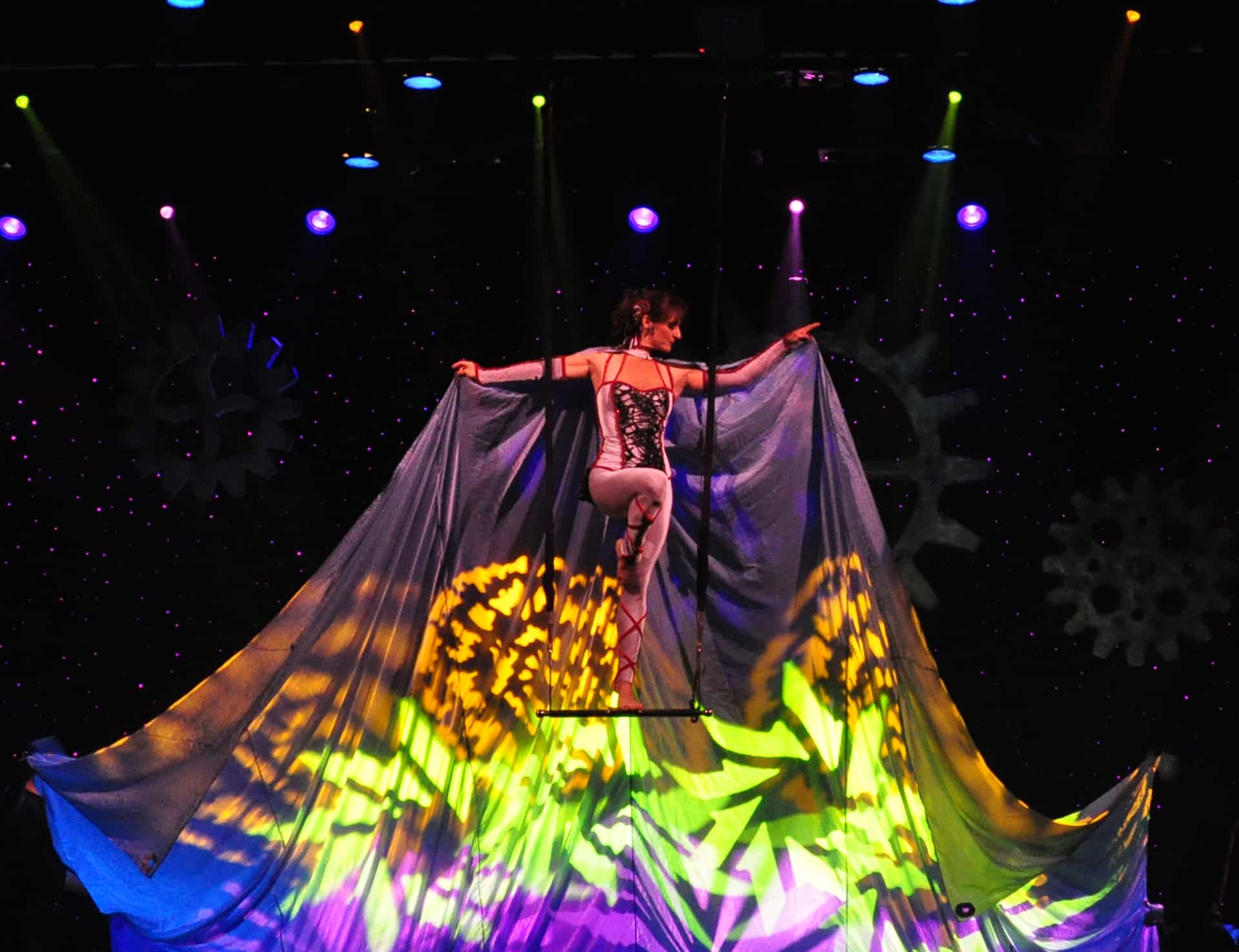 Thin woman standing on a trapeze bar spreading a large colorful cape flowing  behind her
