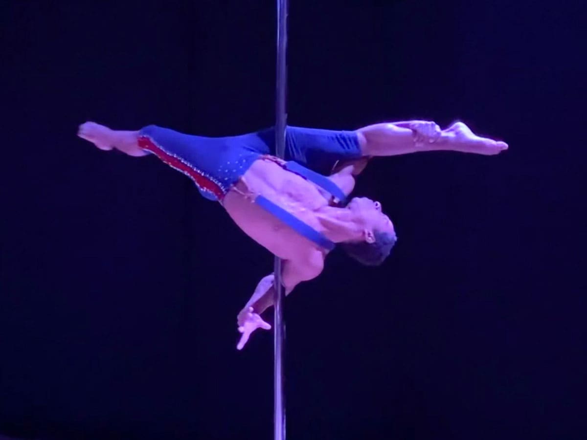 A man wearing blue knee length pants with sparkles and a red stripe down the side along with blue suspenders is spinning around a pole with his body upside down and his legs in a split position.