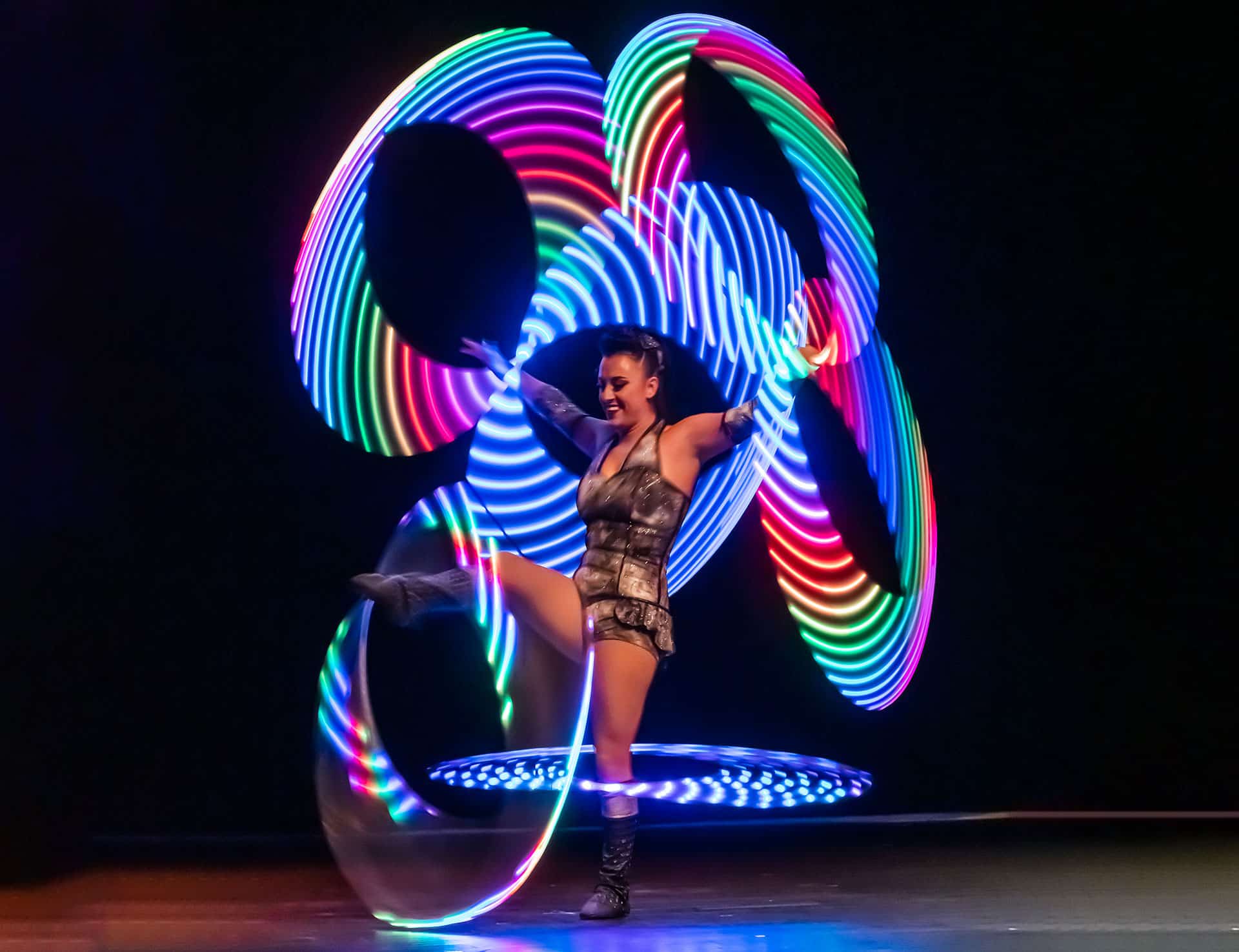 A young woman with dark brown hair standing on her left leg and spinning silver hula hoops on her arms and right leg. She is wearing a black and red costume with black boots and has a backdrop of a circus ring behind her.