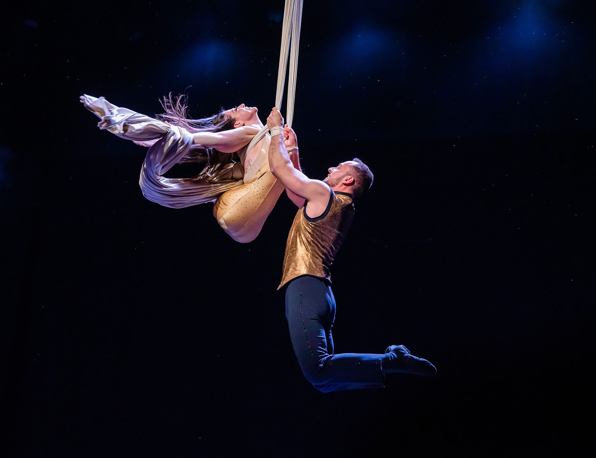 A woman with a flowing cape swinging from a rope steadied by a male partner.