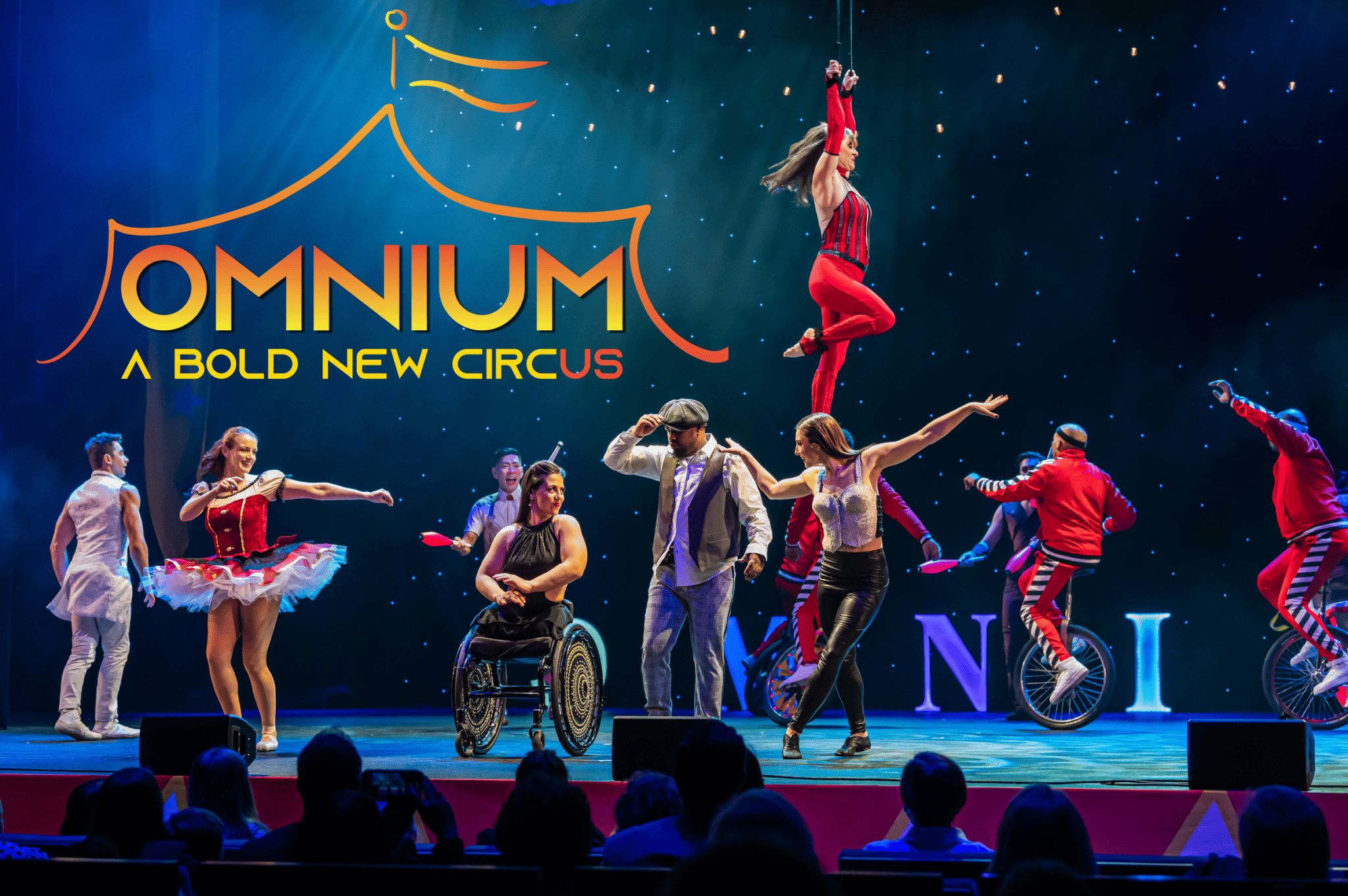 A scene from the opening act of Omnium Circus shows “Johnny” fully immersed in the Circus, standing next to Jen Bricker-Bauer in her wheelchair as multiple dancers dance around them. Vivien hangs in the air while The King Charles Troupe rides their unicycles together in the back. The Omnium Logo is in the top left corner.