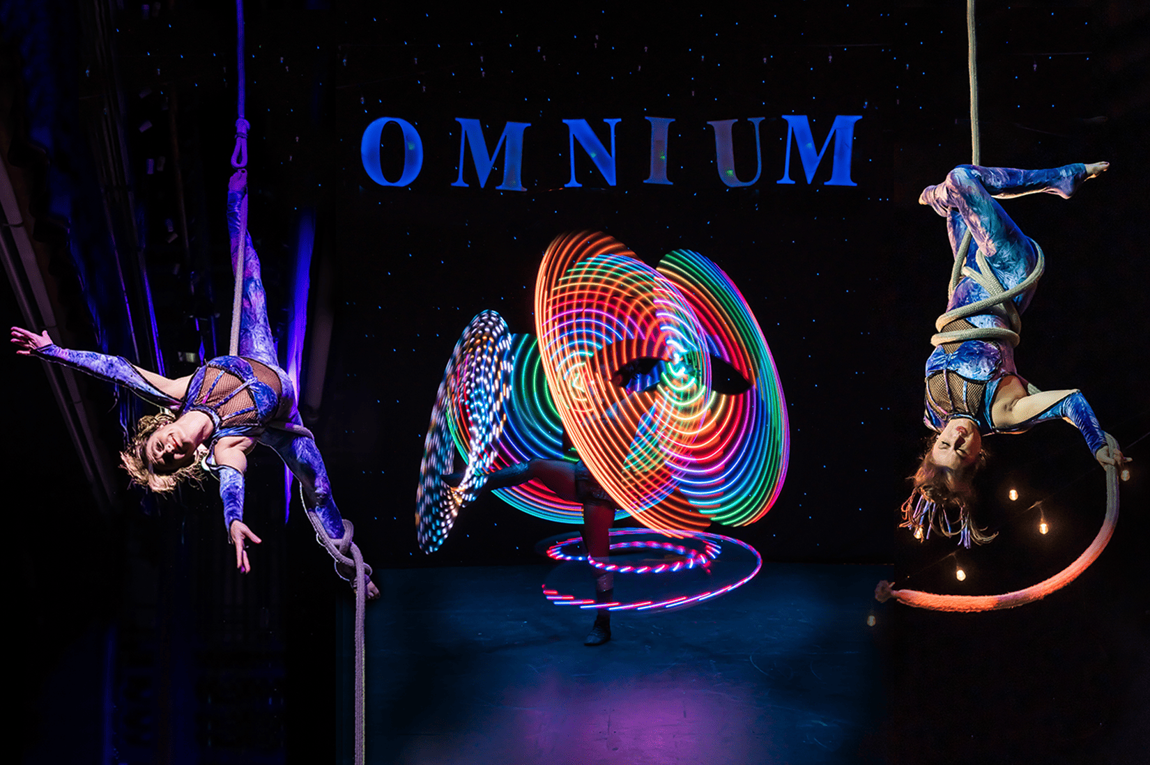 Aerialists perform alongside a woman spinning glow-in-the-dark hoops.